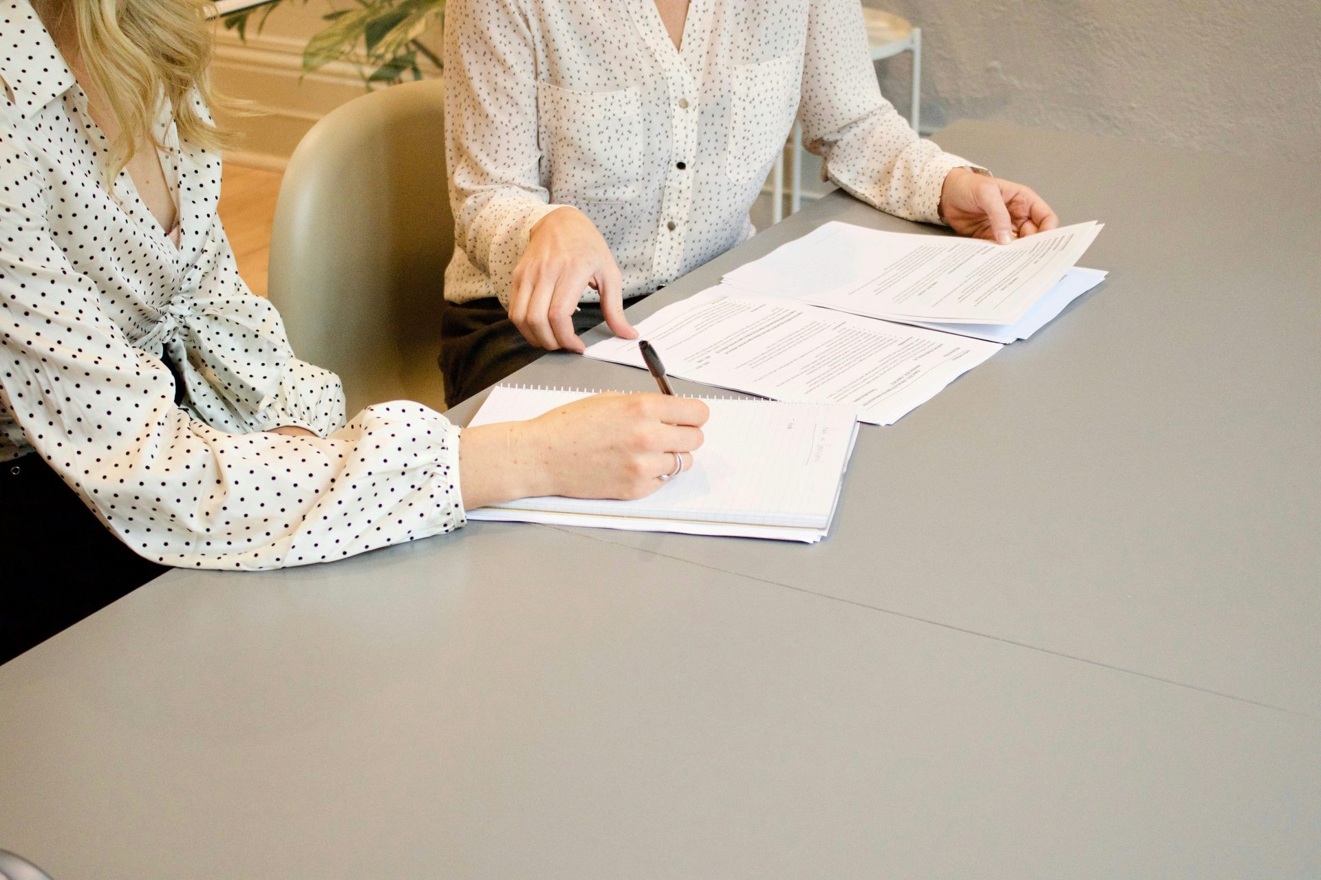 Une femme signe sur une feuille de papier blanc pour imprimante, à côté d'une femme sur le point de toucher les documents.